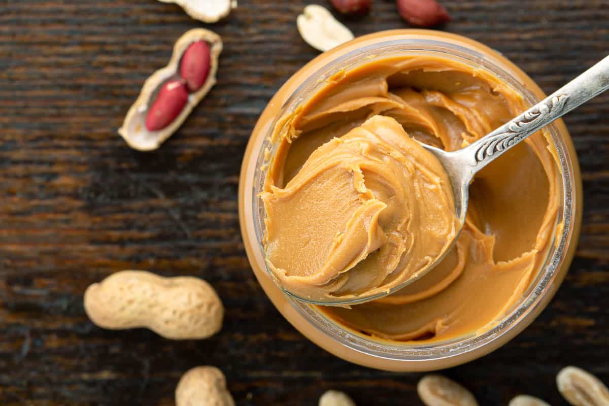 Peanut butter in an open jar and peanuts in the skin are scattered on the table