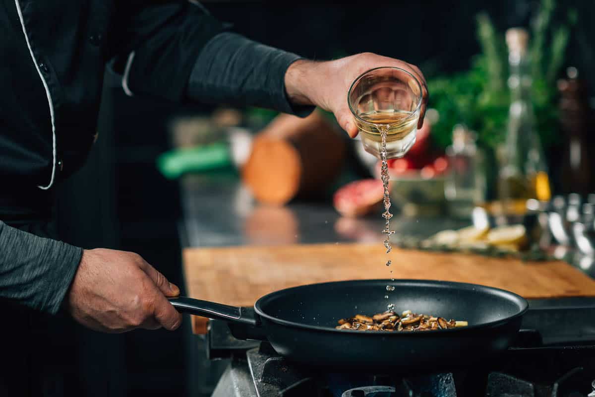 Chef pouring white wine into a frying pan with sliced shiitake mushrooms.