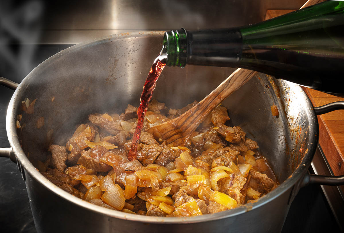 Red wine is poured from a bottle into a pot with roasting goulash to deglaze.