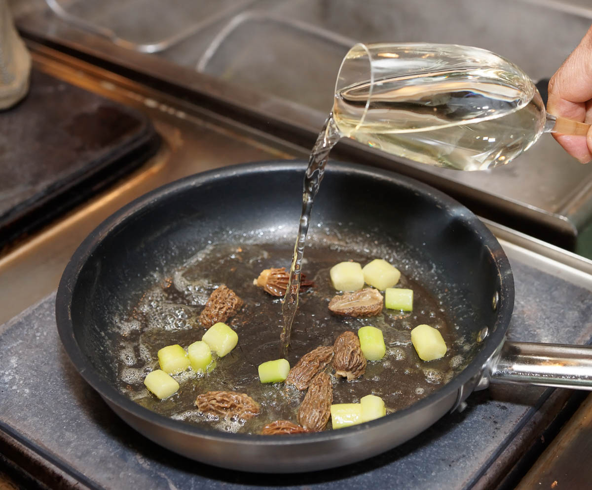 Chef is adding white wine in pan with vegetables and morels.