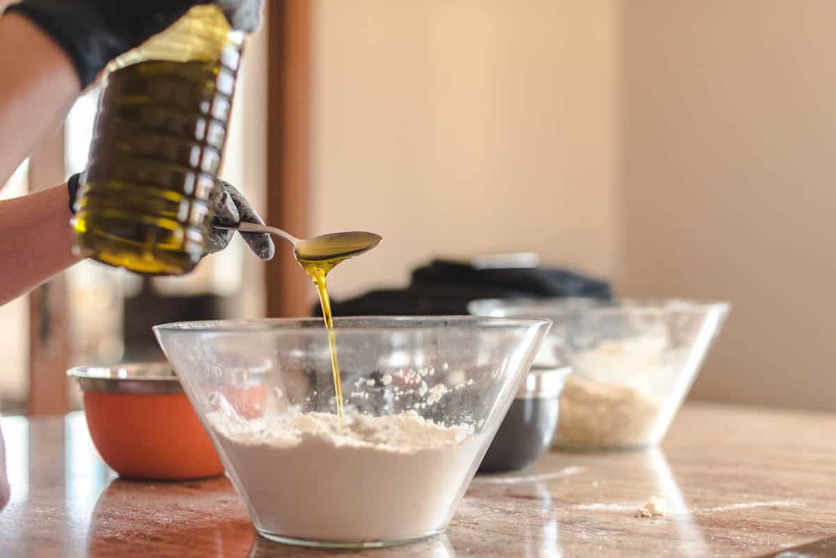 Detail of a young woman pouring olive oil from the bottle into a silver spoon and then into a glass container with flour.