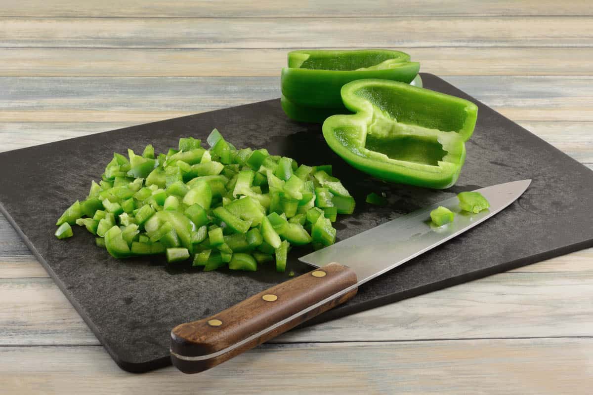 Chopping green bell peppers on black cutting board with kitchen knife.