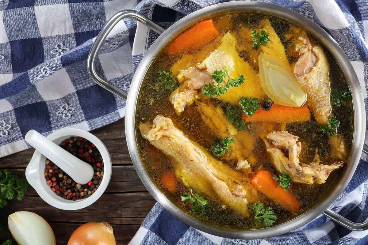 clear Chicken broth with pieces of free range farm organic rooster meat on bone in a metal casserole with kitchen towel and ingredients on wooden table.