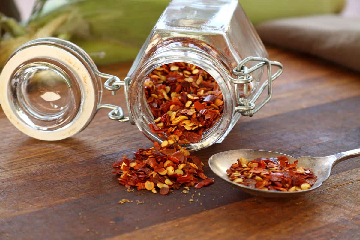 Glass container with dried chili flakes lying on a cutting board.