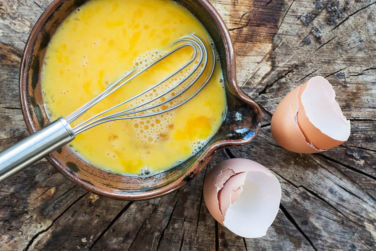 Beaten eggs and whisk in a ceramic bowl. In this photograph the bowl of eggs is placed on a rustic wooden surface, the whisk is placed in the bowl of eggs and is projecting out of the photograph. The egg shells from the beaten eggs are placed on the wooden surface.