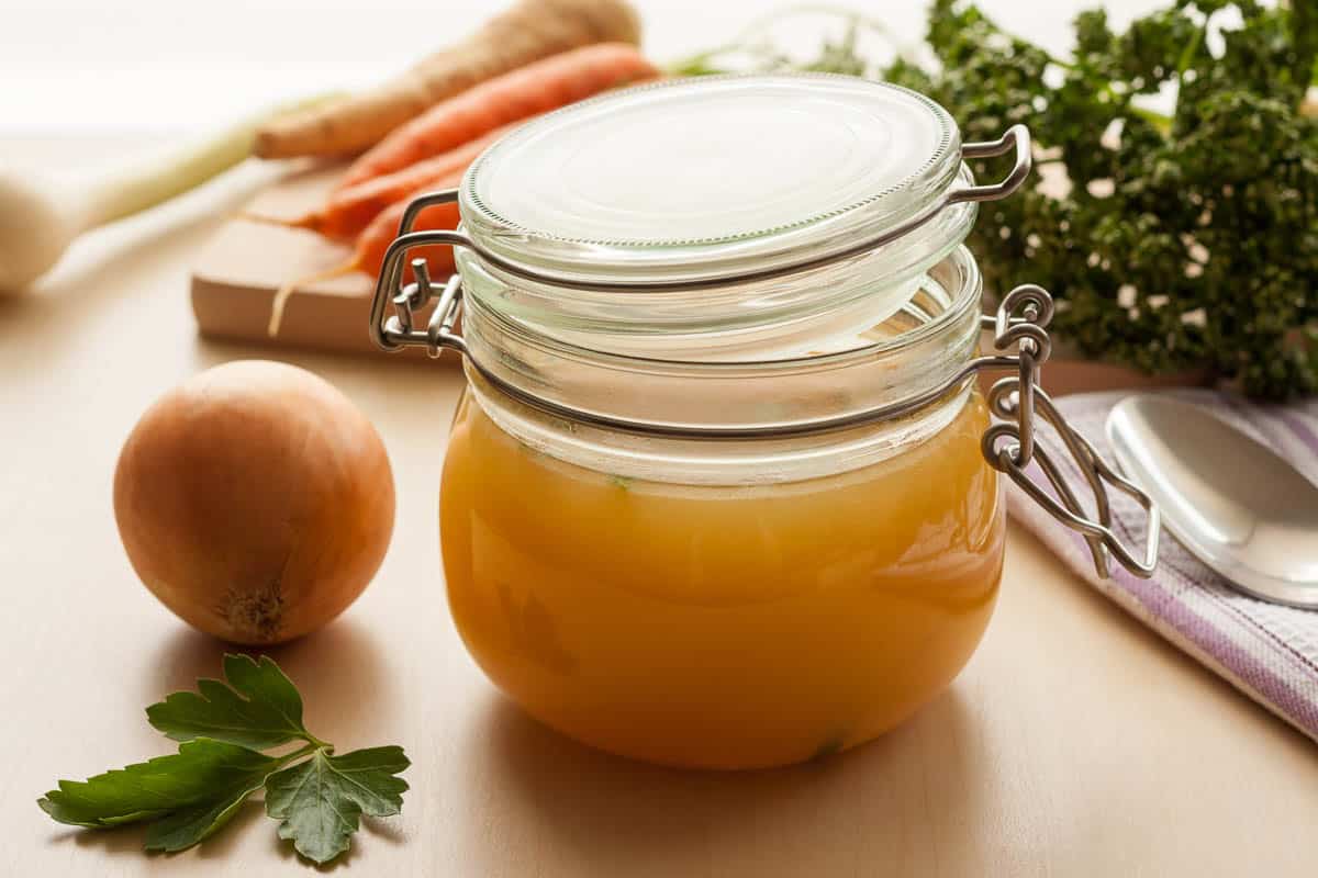 Bone broth made from chicken in a glass jar, with carrots, onions and parsley in the background.