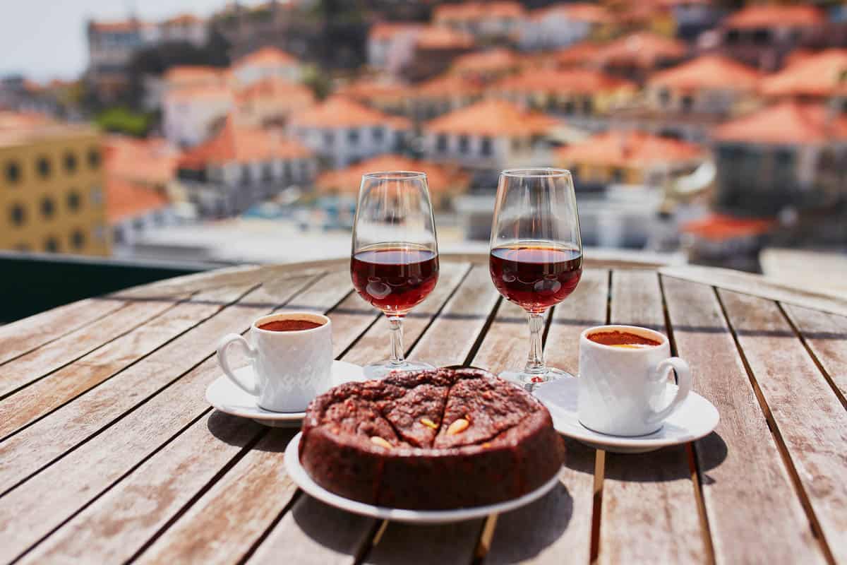 Two glasses of Madeira wine, two cups of fresh espresso coffee and traditional Portuguese honey and nut dessert bolo de mel in cafe with view to Funchal town, Madeira, Portugal.