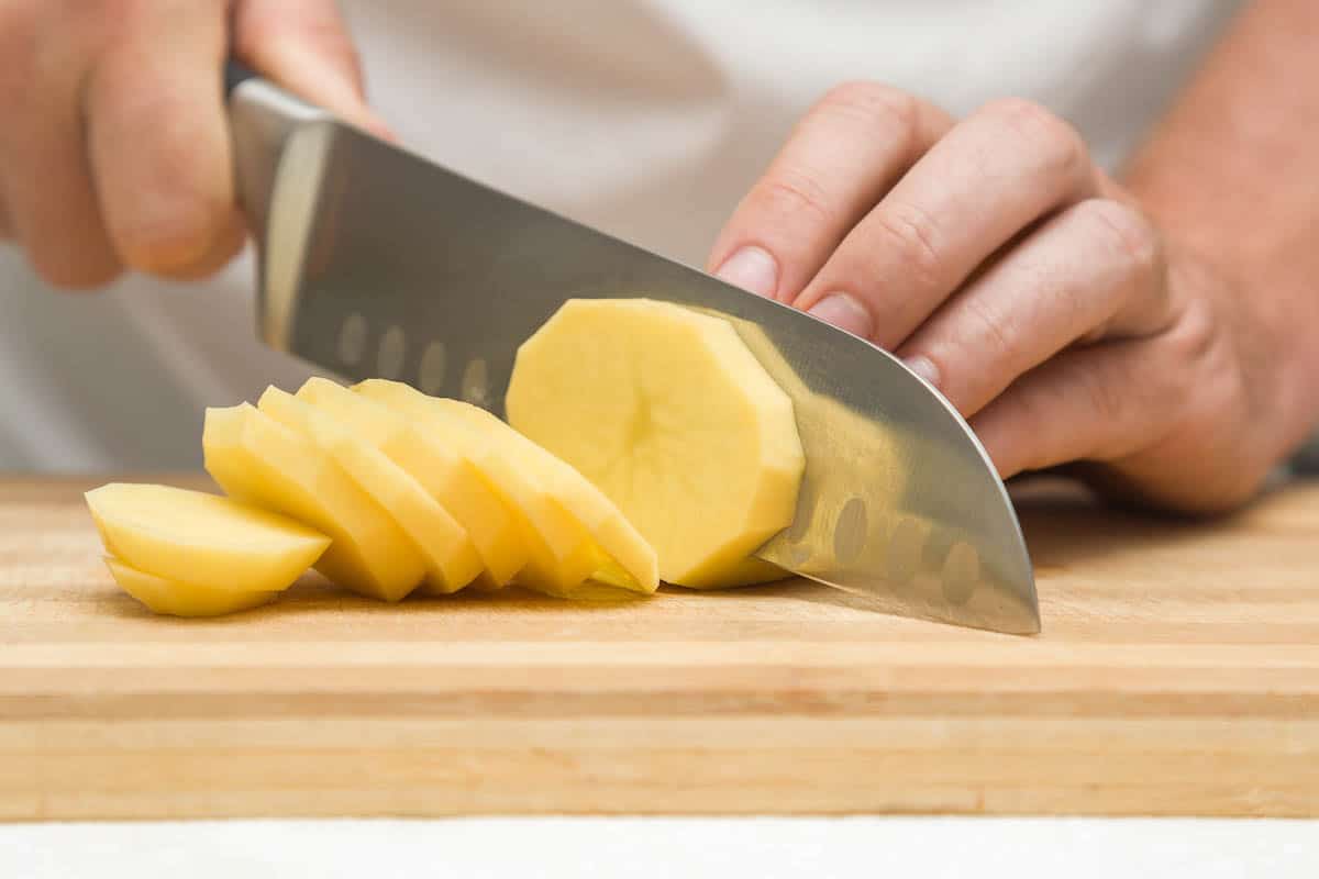 Chef's hands with knife cutting the potato on the wooden board. Preparation for cooking.