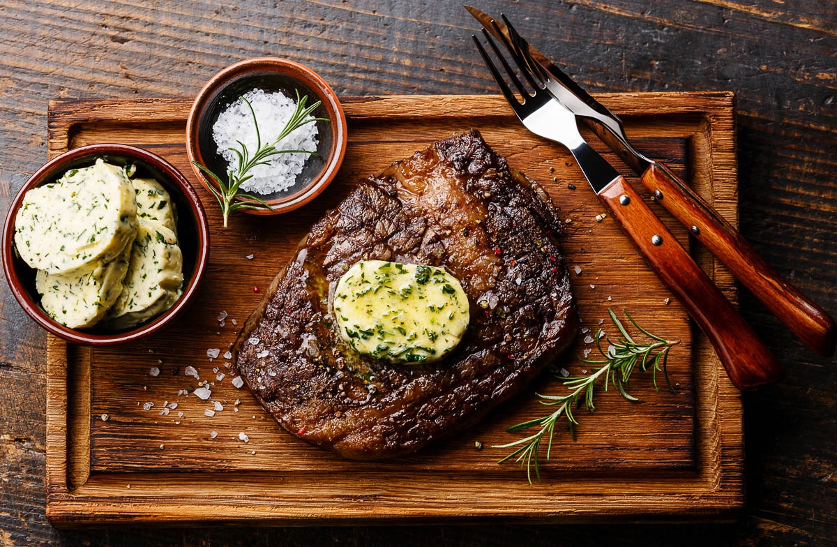 Sliced grilled steak Ribeye with herb butter on cutting board on wooden background.