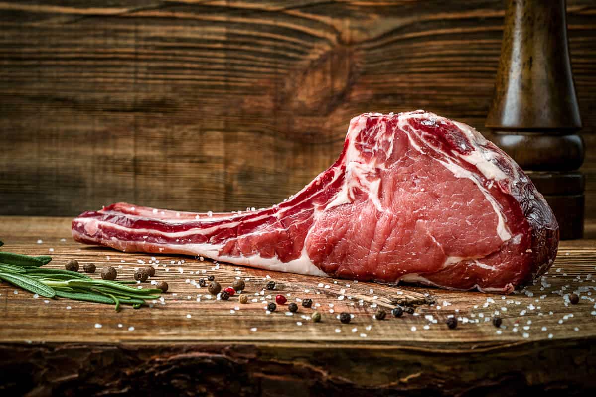Dry aged raw tomahawk beef steak with spices on a wooden background.