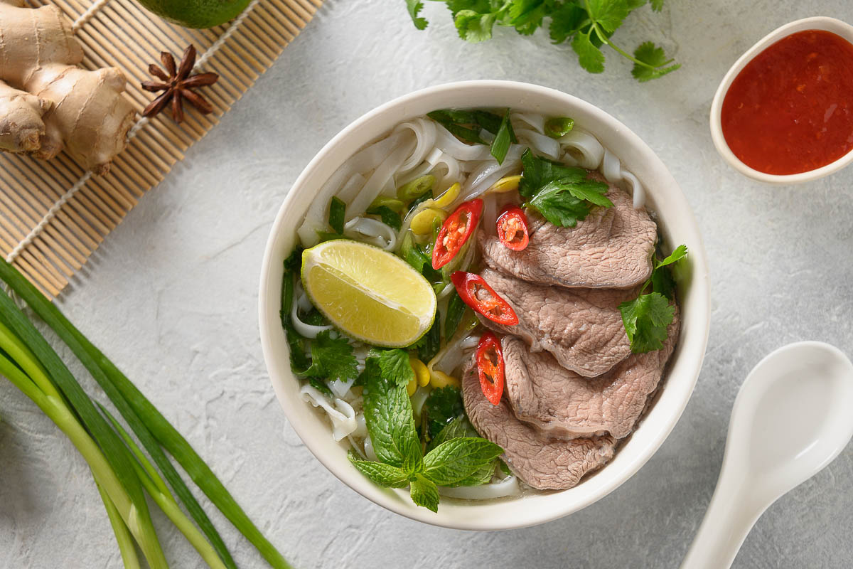 Pho Bo Soup with beef, rice noodles, ginger, lime, chili pepper in white bowl on light backgound.