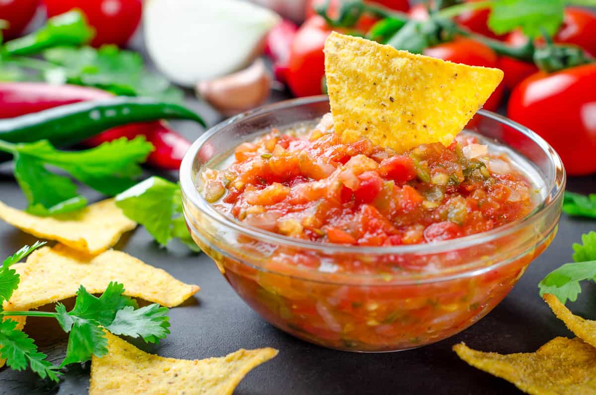 Traditional mexican homemade salsa sauce with ingredients, tomatoes, pepper, cilantro, chips on a dark black table.