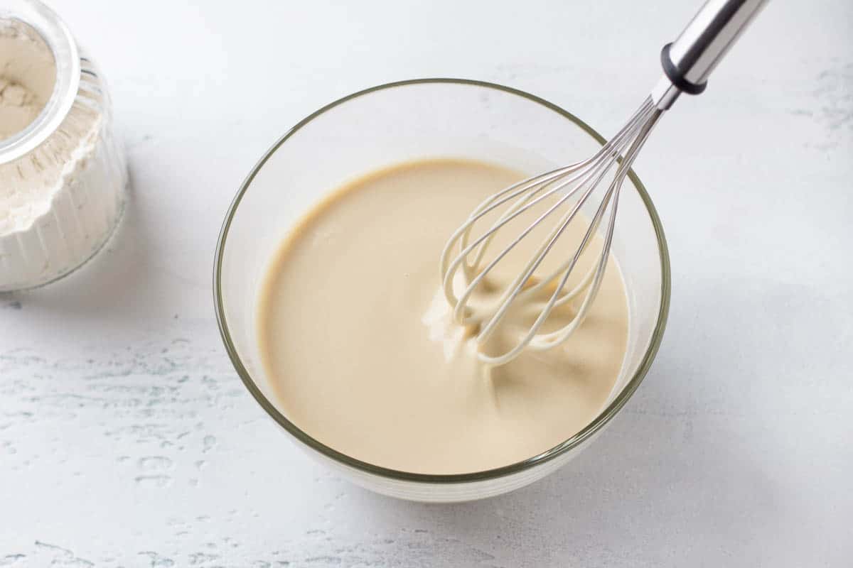 A glass bowl of dough for Finnish pancakes with a whisk and a jar of flour on a light gray background.