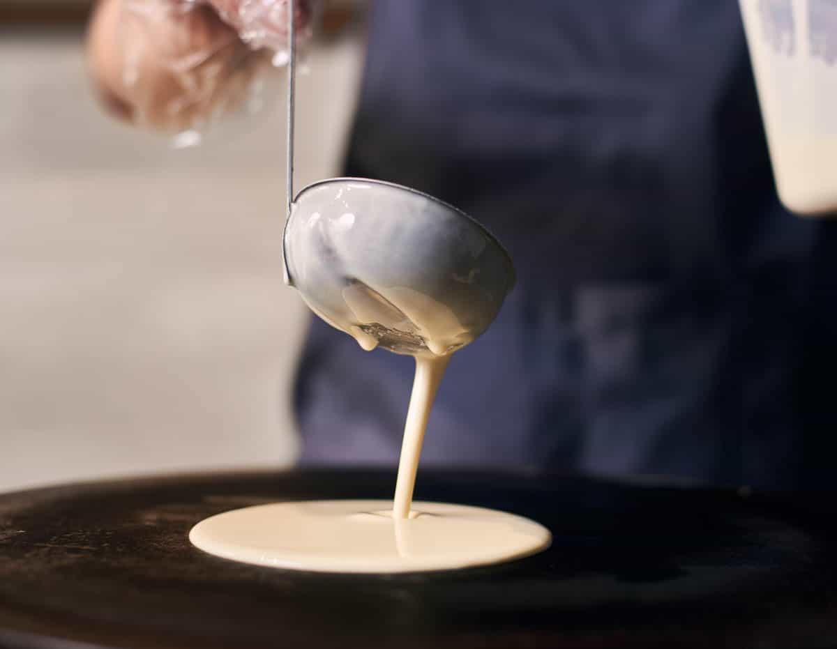 Woman chef in blue apron pour portion of liquid dough by hands in gloves with ladle on electric cooktop for baking pancakes, crepes. 