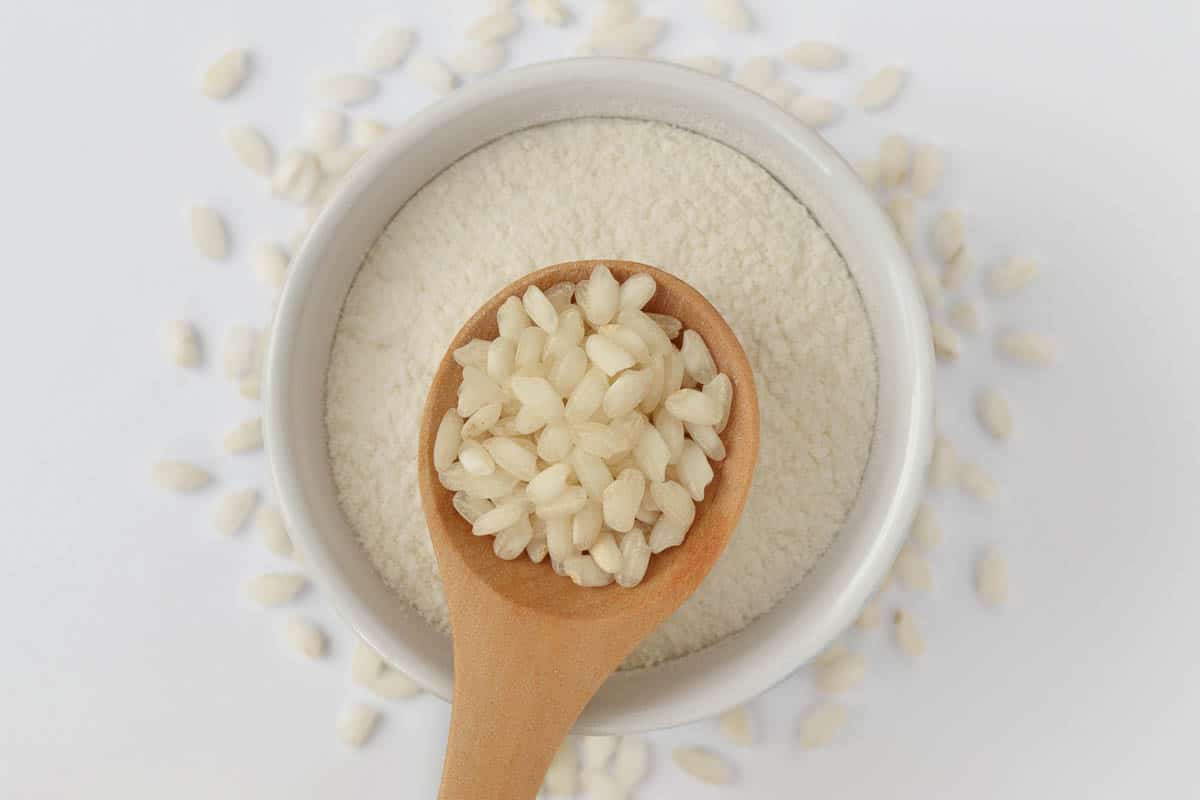 White rice grains on wooden spoon and rice flour in bowl.