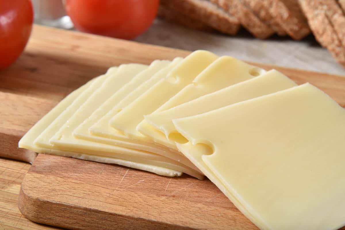 Slices of fresh swiss cheese on a cutting board with tomatoes and whole wheat bread.